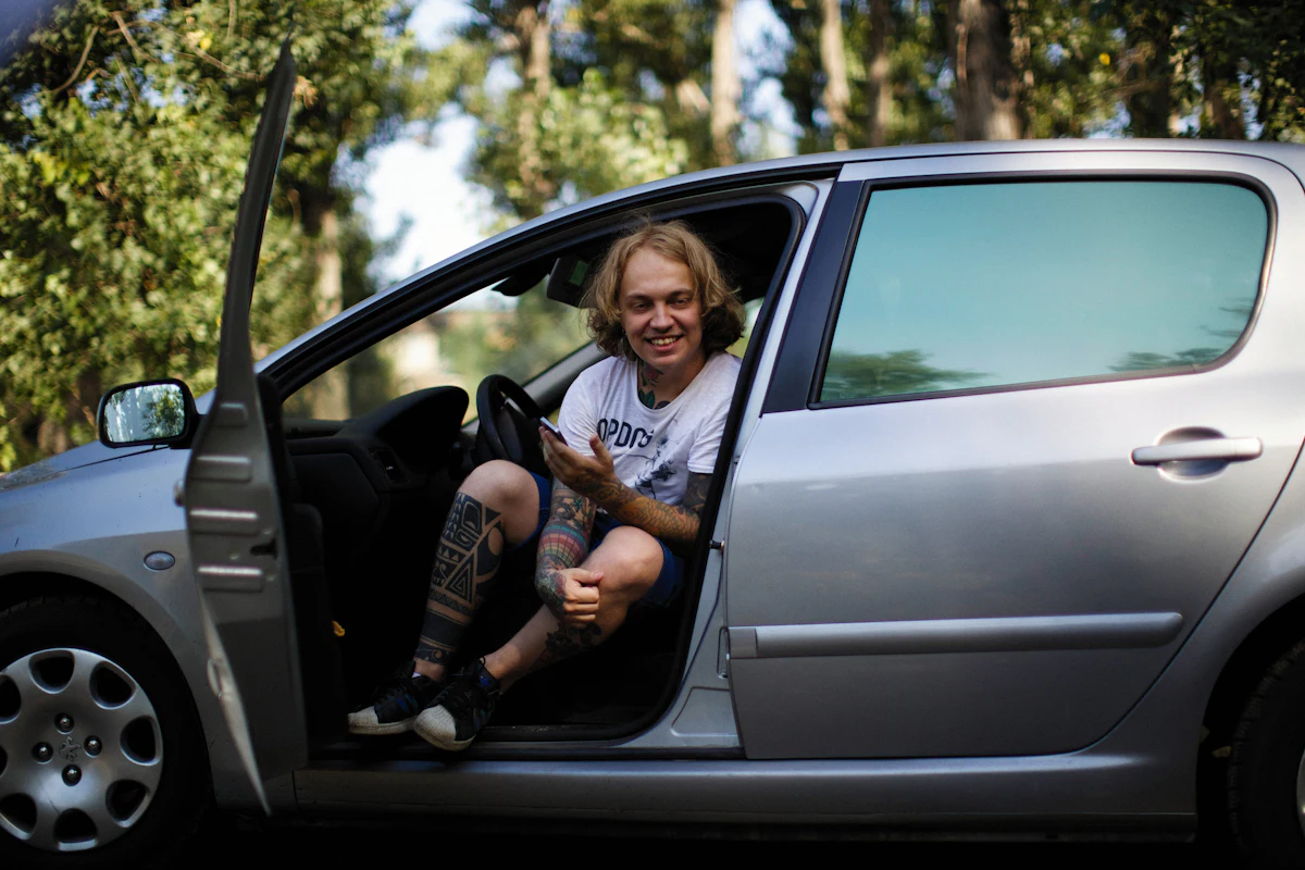 Young man relaxing in the open driver door of a silver used hatchback, smiling