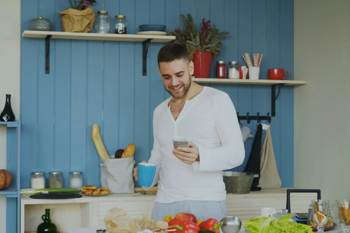 Man smiling while checking his phone with a coffee mug in hand in his kitchen