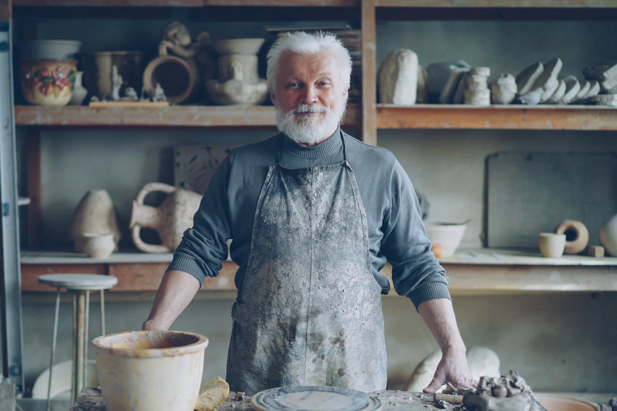 Potter in a work apron standing calmly in his workshop, shelves of finished pieces behind him