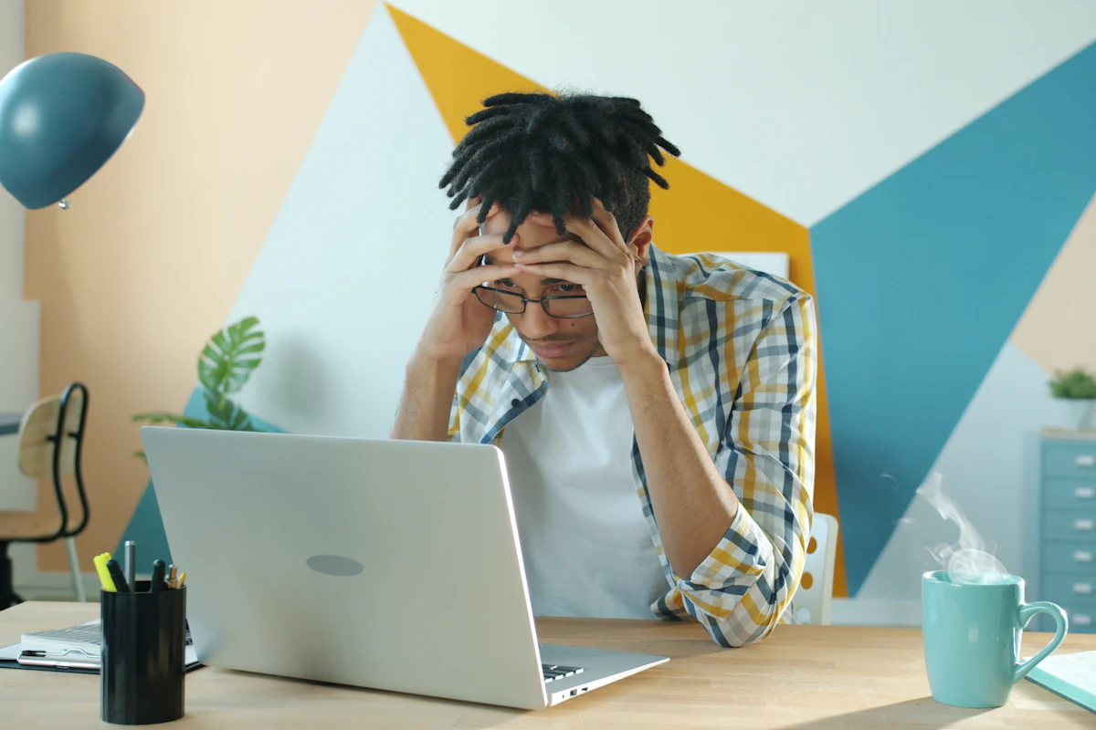 Young business owner at his desk holding his head in frustration in front of a laptop, worried about cash flow
