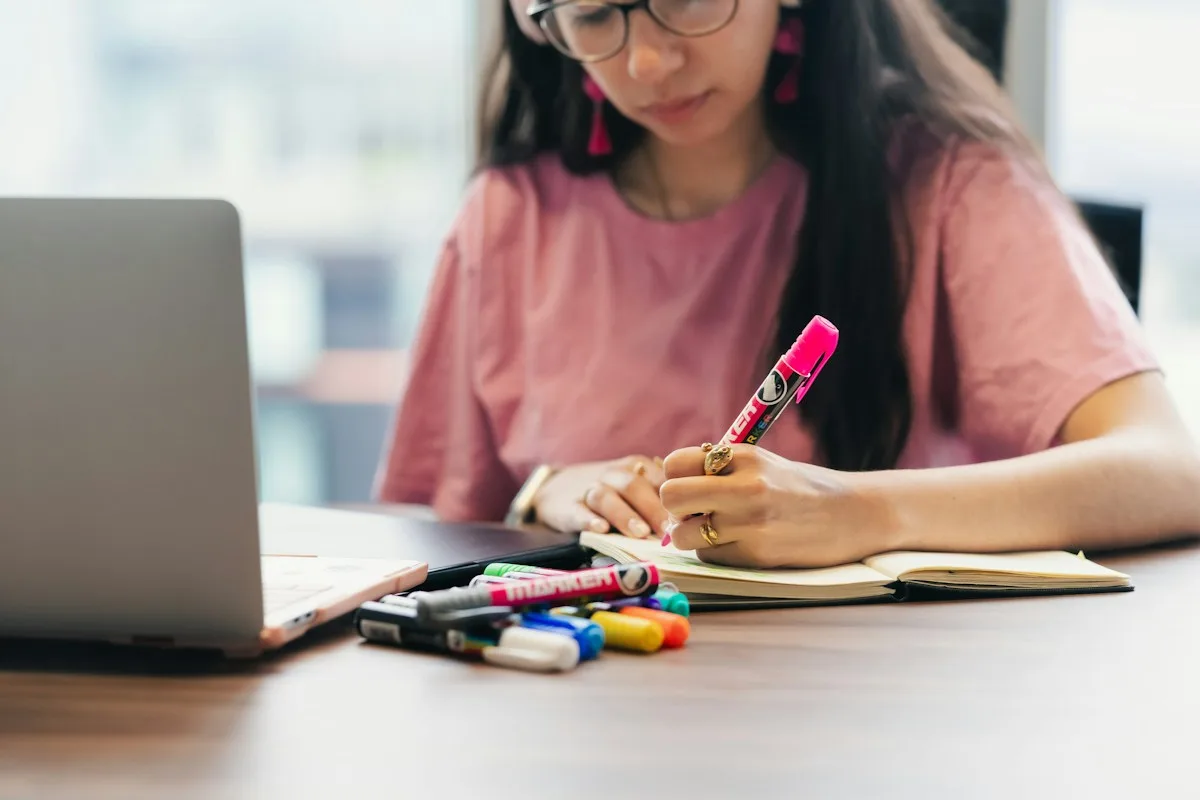 Young woman at a desk writing notes in a notebook next to a laptop, planning her payments